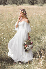 Fototapeta na wymiar beautiful pensive bride in wedding dress and floral wreath holding bouquet of flowers in field