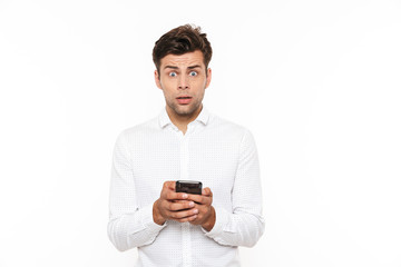 Shocked or outraged man with short dark hair chatting or typing text message using cell phone, isolated over white background