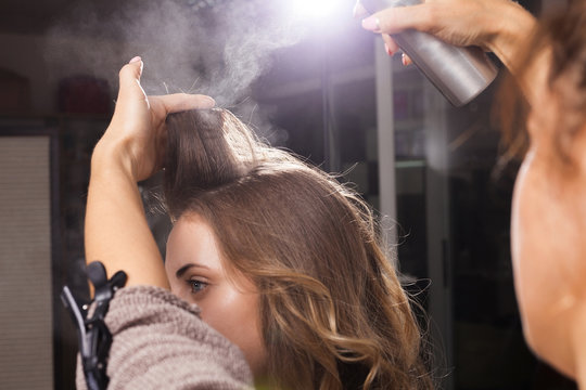 Back view of professional hairdresser fixing a coiffure with curls of a young girl using a hair spray in a beauty salon