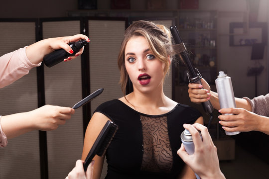 beautiful excited model is surrounded by many hands holding hair curler, sprays and brushes. emotional girl is opening a mouth. concept of professional makeup products