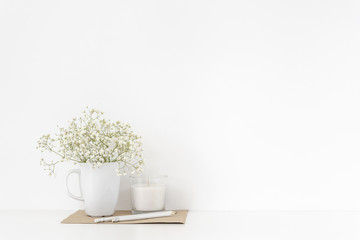 Background with stationary, white candle and bouquet of white flowers in mug on white wall background, elegant soft home decor. Copy space for text. Empty space for lettering, text.