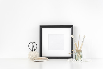 Black square frame mockup in interior. poster mock-Up poster or photo frame and supplies on table near white wall. Hipster minimalism loft desk space, copy space. Background. Back to school.