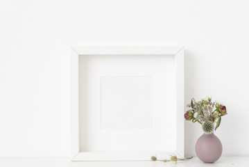 White square portrait frame mockup with dried field wild flowers in vase on white wall background. Empty frame, poster mock up for presentation design. Template frame for text, lettering, modern art.