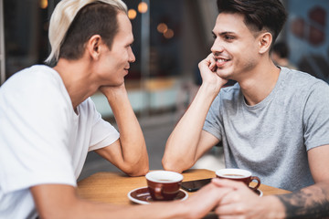 Side view positive man speaking with optimistic friend while drinking mugs of delicious coffee. They sitting at table in cafe