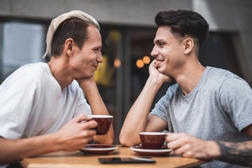 Side view happy man talking with positive friend. They drinking appetizing mugs of liquid while locating at table in cafe and looking on each other
