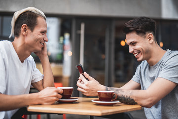 Side view optimistic guys telling together while tasting delicious cups of coffee. They sitting at desk. Positive man using contemporary phone in arms