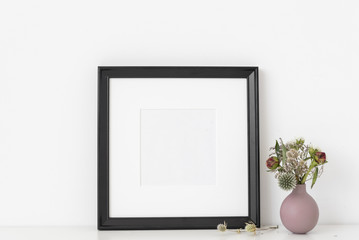 Black square frame mockup with dried flowers in lilac small vase on white wall background. Empty frame, poster mock up for presentation design. Template frame for text, lettering, modern art.