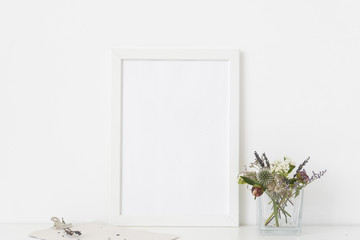 White a4 portrait frame mockup with small bouquet of dried flowers in transparent vase and clamp on white wall background. Empty frame, poster mock up for presentation design. 