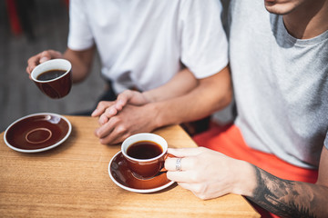 One guy holding hand of another man. They sitting at table and drinking cups of appetizing coffee