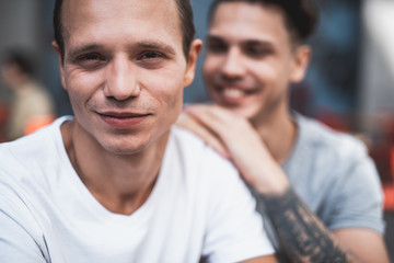 Optimistic man looking at camera while putting hands on shoulder of smiling comrade during conversation with him. Glad friends spending time together concept