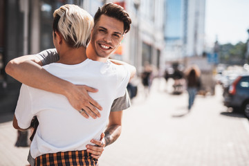 I am so happy to see you. Beaming boy embracing friend while locating at street on sidewalk. Different people walking there