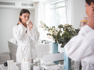 Portrait of calm girl with closing eyes tasting appetizing mug of liquid while wearing bathrobe after taking bath. She reflecting in mirror in room