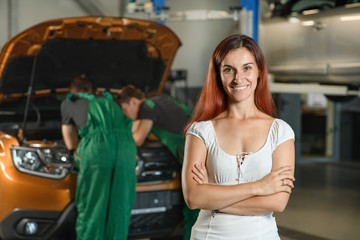 A beautiful girl poses for a photo while, two young mechanics in green overalls try to fix the engine of an orange car