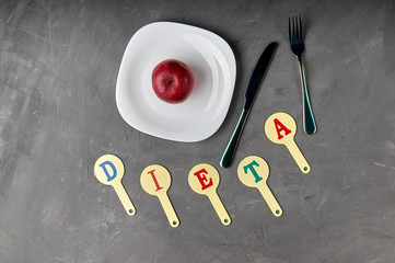 The word "diet" is collected from a children's educational card with letters. Unusual black cutlery, fork and knife ready for breakfast, lunch, dinner. One ripe red apple on a plate, too little food.