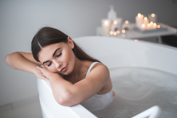 Portrait of calm woman with closing eyes enjoying during cosmetic procedure in contemporary bath. Candles locating opposite her. Pleased lady taking care of body during romantic atmosphere concept
