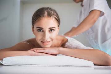 Obraz na płótnie Canvas Enjoying spa. Close up portrait of charming girl looking at camera with smile while having spa procedure. Masseur doing back massage on blurred background