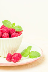Ripe raspberries with green mint leaves in cup and saucer on pastel yellow background.