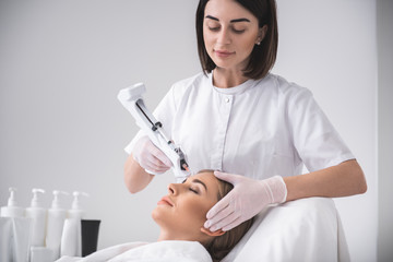Helpful procedure. Attractive young woman with closed eyes relaxing at beauty salon. Cosmetologist using automatic syringe gun