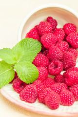 Ripe raspberries with green mint leaves in cup and saucer on pastel yellow background.