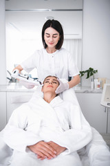 Skincare treatment. Attractive serene woman laying at daybed with closed eyes while having beauty procedure. Cosmetologist in lab coat using electronic syringe gun