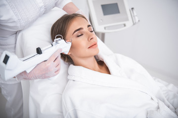 Day off at spa. Smiling girl with closed eyes lying on daybed while having beauty procedure. Cosmetologist hand in sterile glove holding automatic syringe gun