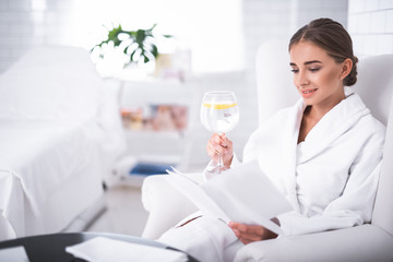 Perfect vacation. Lovely girl in soft bathrobe with papers and refreshing drink relaxing at wellness center