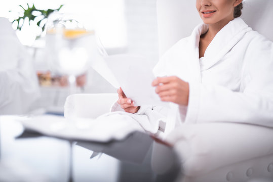 Choosing procedure. Cropped lovely lady in white bathrobe sitting on armchair and holding booklet