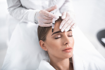 Rest and care. Close up portrait of attractive girl with closed eyes and cosmetologist hands in gloves. Doctor touching lady forehead and using syringe © Yakobchuk Olena
