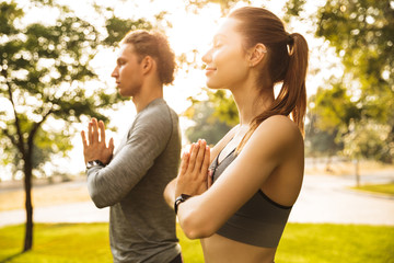 Image of athletic sporty couple man and woman 20s in tracksuits, holding palms together while working out or doing yoga in green park