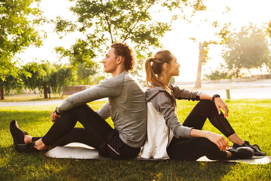 Image of young sporty couple man and woman 20s in tracksuits, sitting on grass in green park back to back