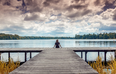 Girl woman sits with her back on a wooden pier and looks at the landscape, lake and clouds, Trakai, Lithuania