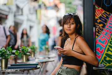 A young Asian Chinese teenager sitting outside the cafe while holding her smartphone. She is...