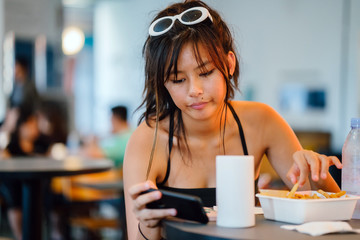 A young millenial Chinese Asian is having some snacks while reading through her smartphone. She looks preoccupied and quite bored.