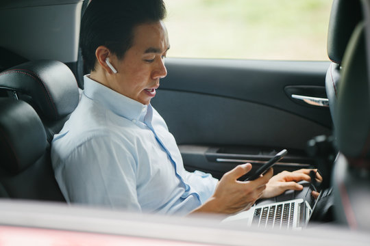 A Young Japanese Asian Man Is Talking On His Smartphone Using His Bluetooth Headset. He Is Riding At The Back Seat Of A Car He Booked Using A Ride Hailing App.