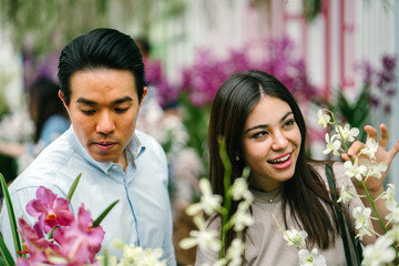 A young and attractive Asian couple stop to smell the flowers in a park during the day. They are both good looking and are smiling as they enjoy a sunny day outside sightseeing together.