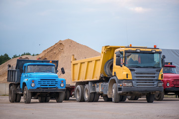 Multiple cars, trucks, loaders, concrete mixers and construction machinery in large parking lot in industrial territory, next to concrete and asphalt factory. Raw material heaps, gravel in background