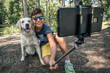 A young boy and his dog taking a self portrait with selfie stick and trees in background. A smiling teen make selfie with a doggie on a smart phone outside in the park. Teen taking a self picture