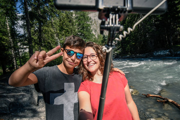 Couple of friends taking self portrait with selfie stick with a mountain river in background. Two young brothers make selfie on a phone outside in the park. Teens taking a self picture with smartphone