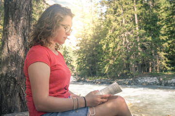 Teen relaxing reading a book near the river with a mountain forest in background. Pretty relaxed young woman read a book outdoor in the nature with sun shining. Portrait of a cute girl under tree.