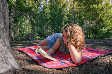 Teen relaxing reading a book lying on a plaid with a mountain forest in background. Pretty relaxed young woman on a rug read a book outdoor in the nature. Portrait of a cute girl with mini skirt.