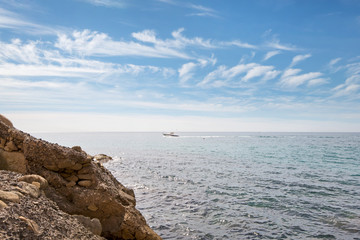 Rocky mountain coastline where rocks meet the sea, close to a busy harbor in a port city. International trade ships and recreational vessels can be found close to the shore. Sailing yachts and boats