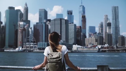 Back view of young happy tourist girl with backpack and arms wide open with joy at famous Manhattan skyline in New York