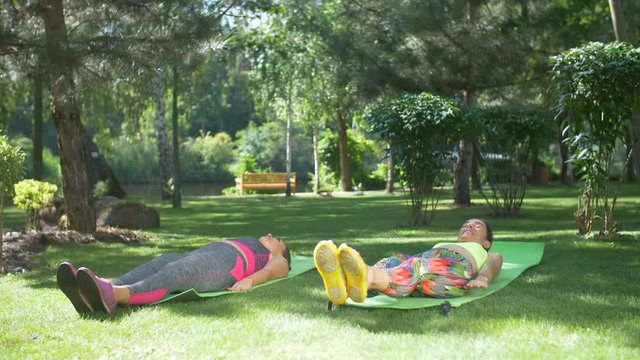 Concentrated fitness women in sportswear practicing v-ups crunches ab toning exercise workout in summer park. Smiling fit females working out outdoors doing sit-ups exercise for upper and lower abs.