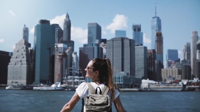 Camera follows young happy traveler girl with backpack, jumping with arms wide open at Manhattan skyline in New York.