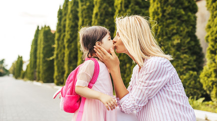 Horizontal portrait of happy cute little girl and beautiful mother embracing and kissing each other on sidewalkbefore the kindergraten. Mom with kid preschooler outdoor. Mother's day, school concept.