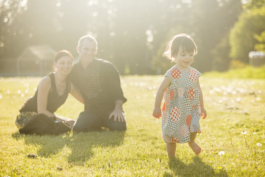 Cute little girl in a dress playing in the grass at sunset with her parents in the background.