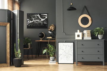 Black painting on grey wall in dark living room interior with plants and poster. Real photo