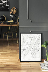 Poster and plant on wooden floor in grey workspace interior with gold chair at desk. Real photo