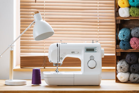 Close-up of a white sewing machine with a purple thread and crates with yarn by a window in a bright crafts room interior. Real photo.