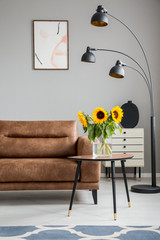 Sunflowers on wooden table next to brown sofa and black lamp in flat interior with poster. Real photo